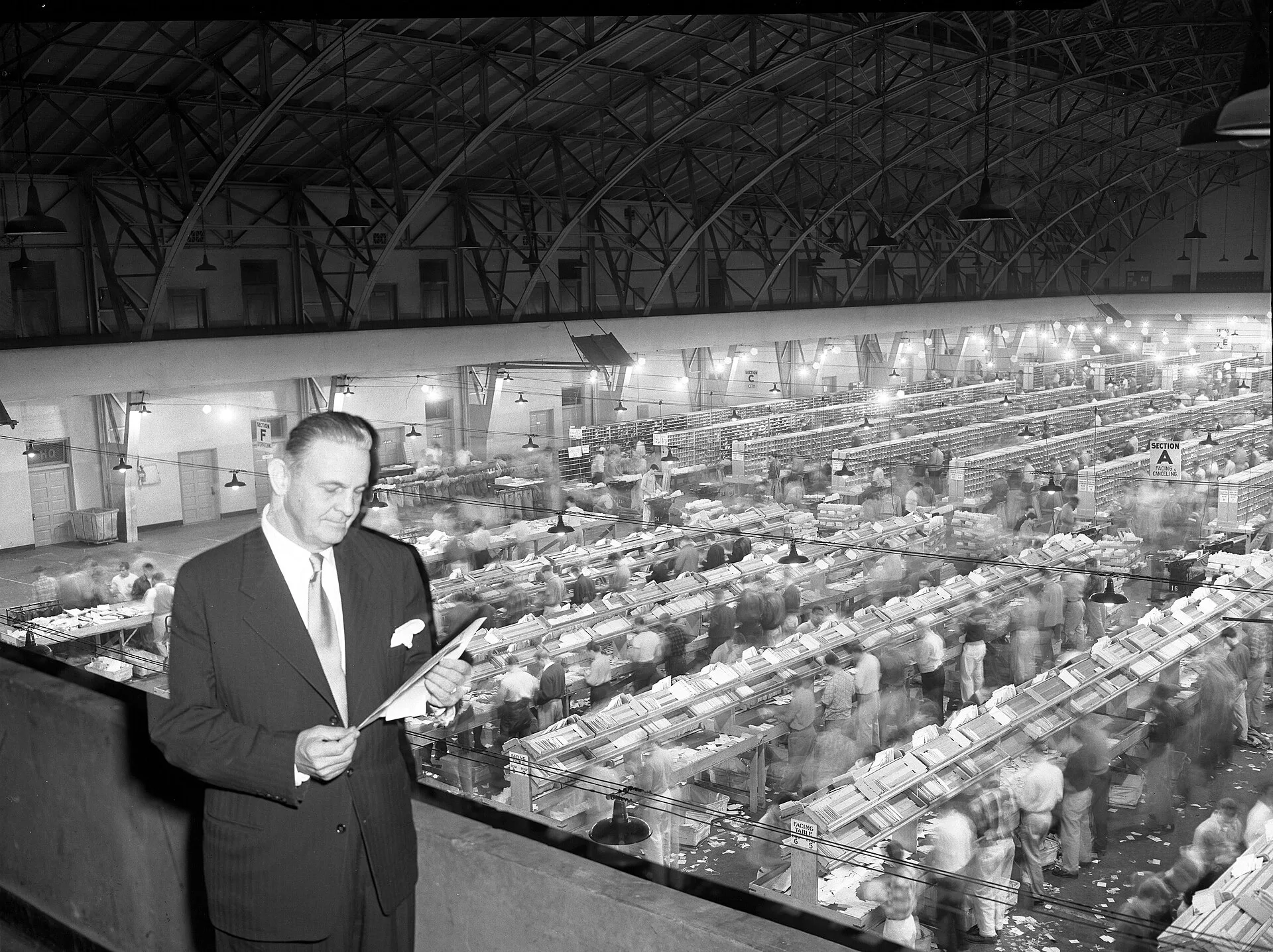 Title: Michael D. Fanning, U.S. Postmaster, standing in rafters above employees working in mailing sorting warehouse in Los Angeles, Calif., 1951. The offline marketing benchmark card that keeps figures checkable